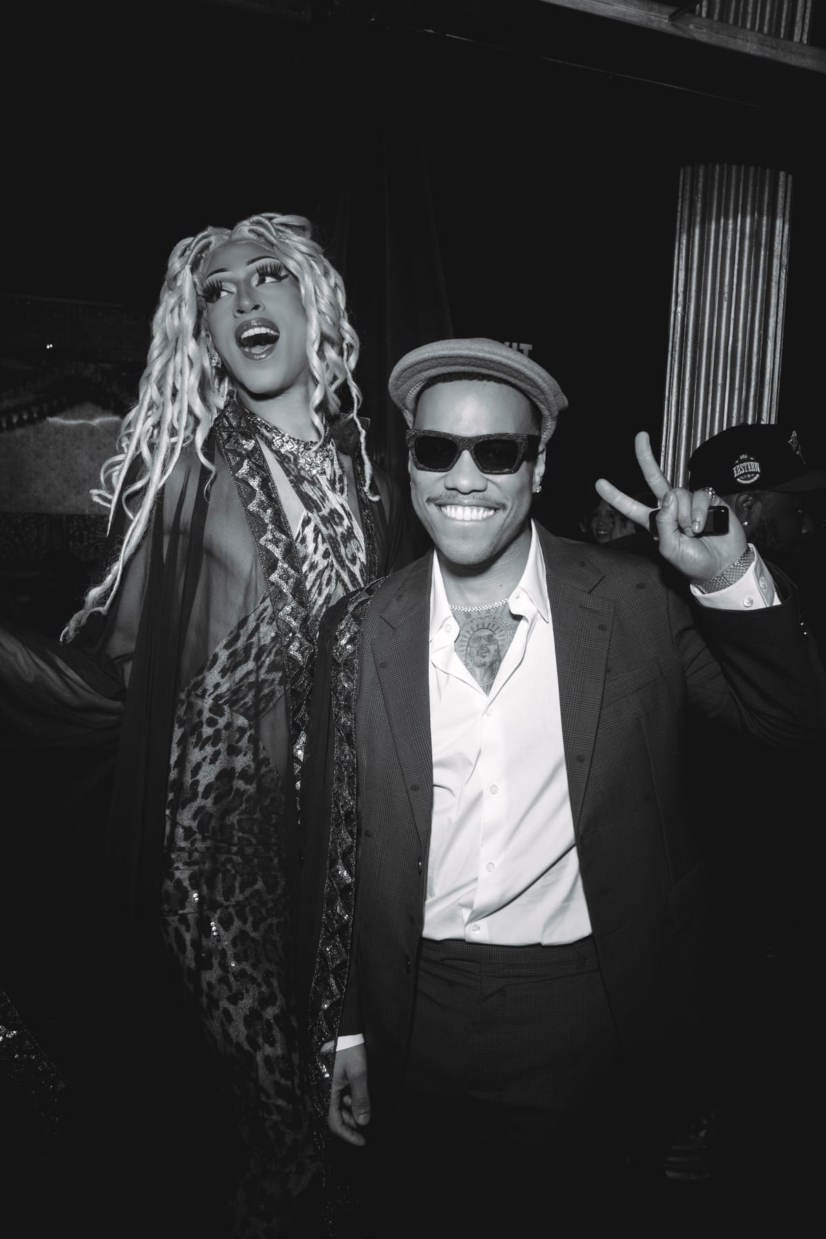 Anderson .Paak in a cap and sunglasses, smiling and throwing a peace sign beside a performer in a leopard-print gown.