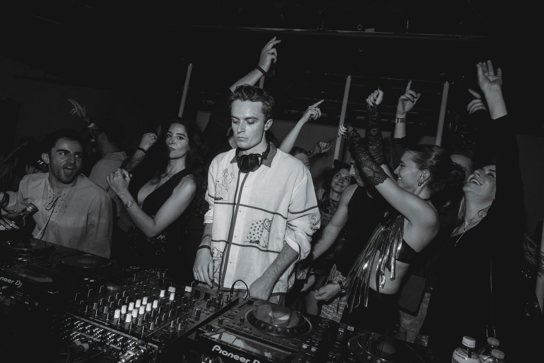 A DJ in headphones and an illustrated shirt at the mixer, with dancers reaching up all around him.