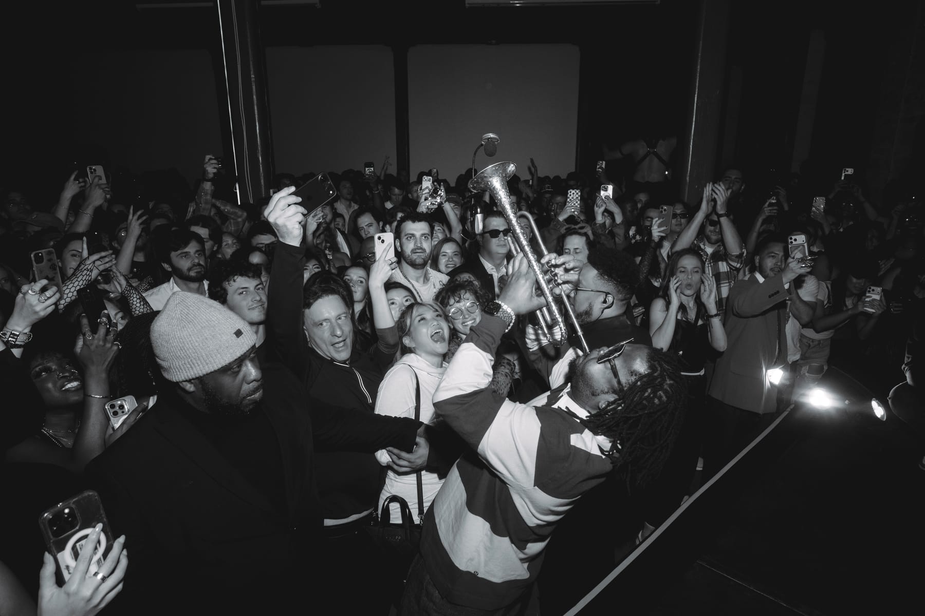 A packed crowd with phones raised around a trumpet player in a striped rugby shirt.