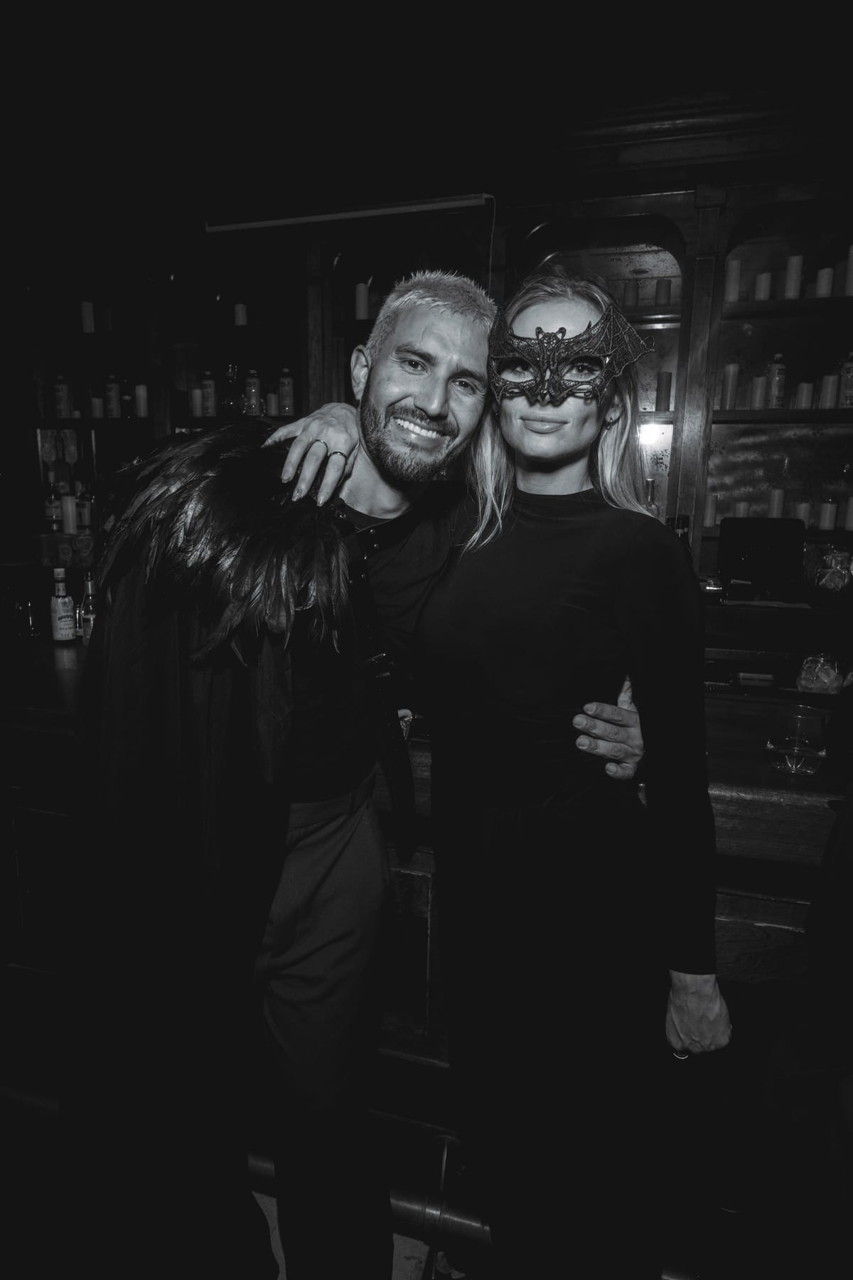 A smiling man in a black feathered cape with his arm around a woman in a lace bat mask.