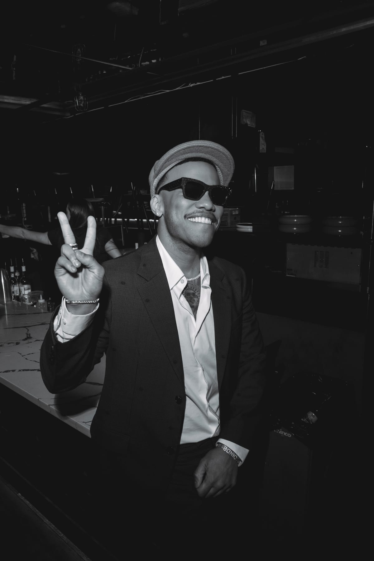 Anderson .Paak in a cap, sunglasses and a suit, smiling and flashing a peace sign at the bar.