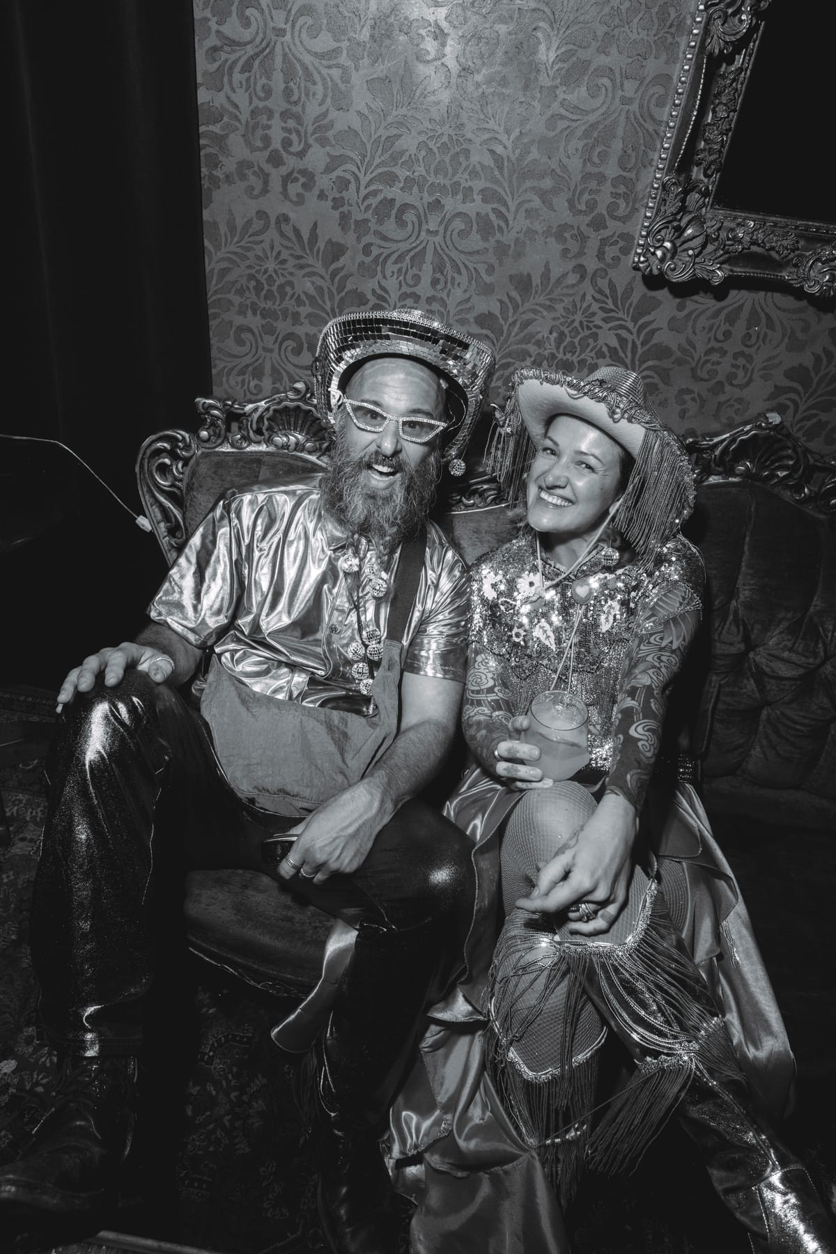 A man in a mirror-ball hat and silver shirt beside a woman in a fringed cowboy hat on an ornate sofa.