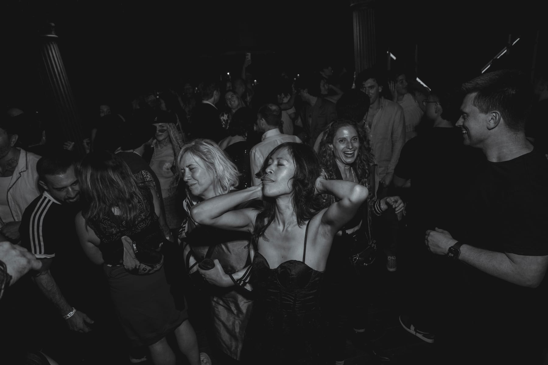 A woman dancing with her eyes closed and hands behind her head, surrounded by a smiling crowd.
