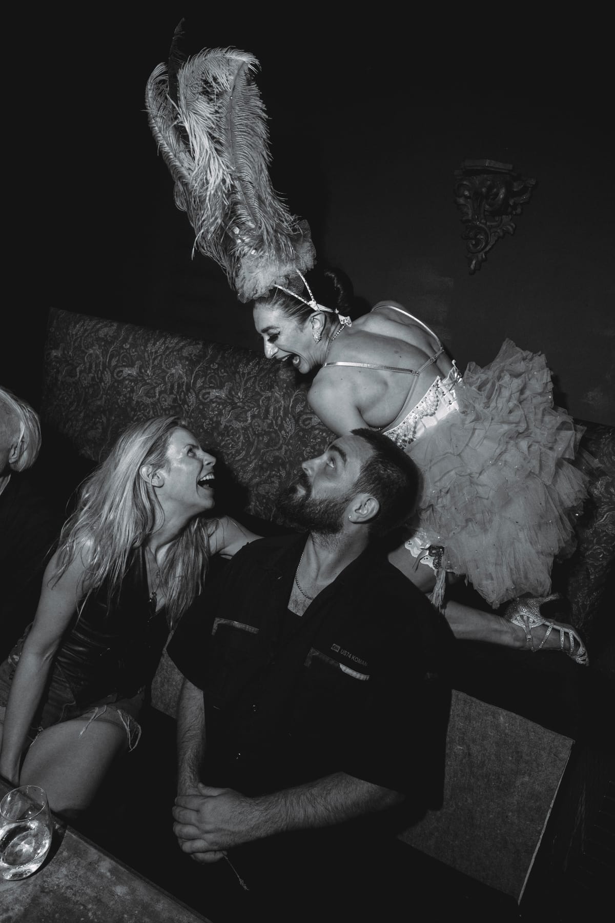 A showgirl in a plumed headdress and tulle leans over a laughing couple on a banquette.
