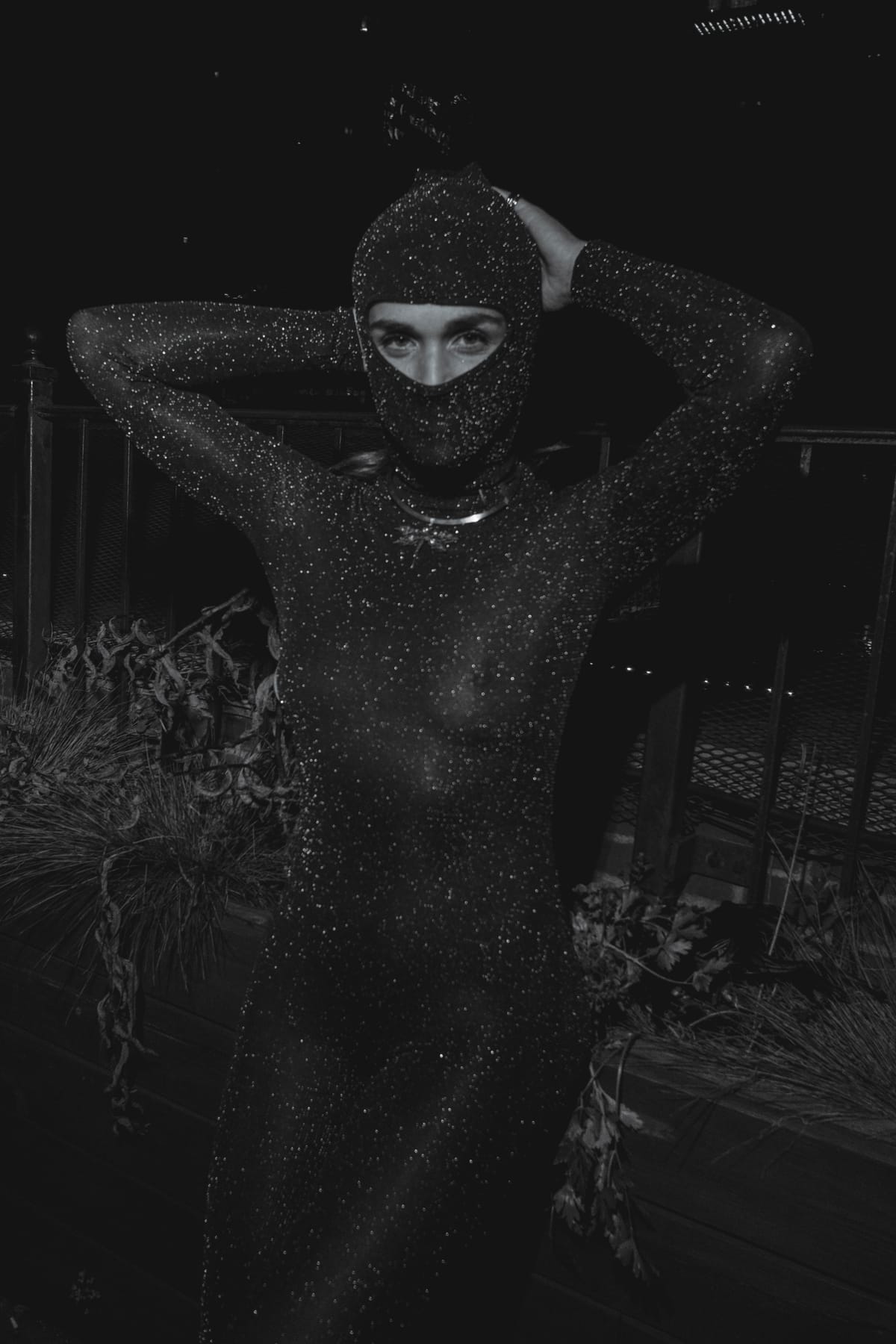 A figure in a sparkling full-body suit and matching balaclava, hands behind the head.
