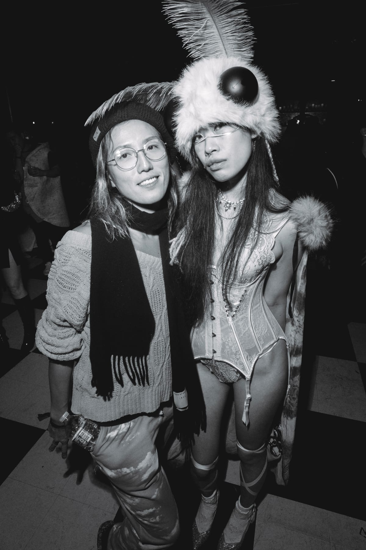 Two friends posing, one in a beanie and scarf, the other in a fluffy moth headpiece and white lace corset.