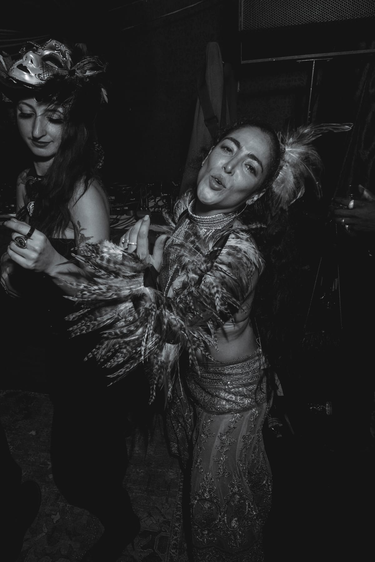 Two women dancing, one in a feathered jacket and headpiece, the other with a mask pushed up on her head.