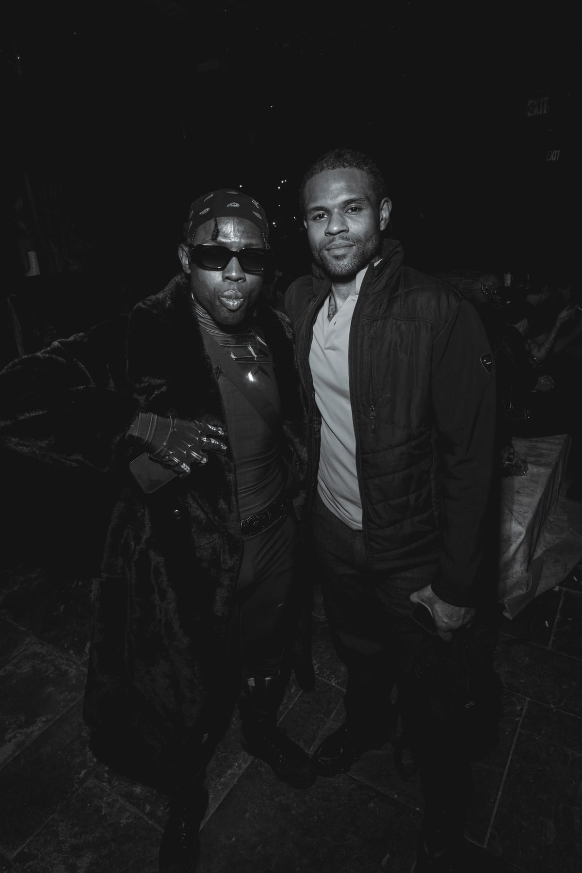 A man in a long fur coat, bandana and sunglasses pouting beside a friend in a dark jacket.