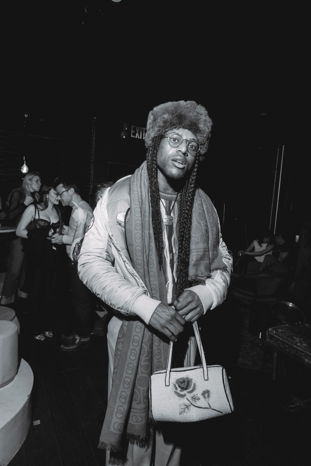 A man in a fur hat, round glasses and a long patterned scarf holding an embroidered handbag.